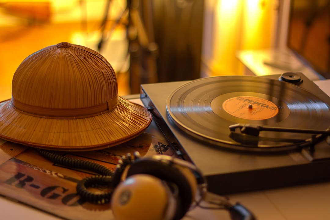 Retro Reverie – Vintage Turntable and R.GO’s Safari Hat in Sunlit Still Life An 1980s Hungarian R.GO vinyl record leans against a retro turntable, its cover’s bold typography and the band’s iconic safari hat draped casually beside it. Summer light slants through half-drawn blinds, striping the scene with golden rays that catch the dust motes and the record’s glossy sheen. The needle hovers just above the vinyl, frozen in a moment between tracks—a tribute to analog nostalgia and Hungary’s pop legacy. by Headphone Brands A record player and safari hat.